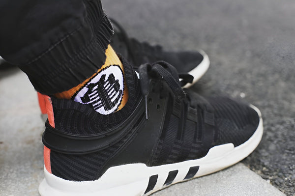 Une personne porte des chaussettes noires avec un motif orange et blanc de Son Goku, un personnage de Dragon Ball Z. Elle porte également des chaussures de sport noires et grises.,CH-0935396012