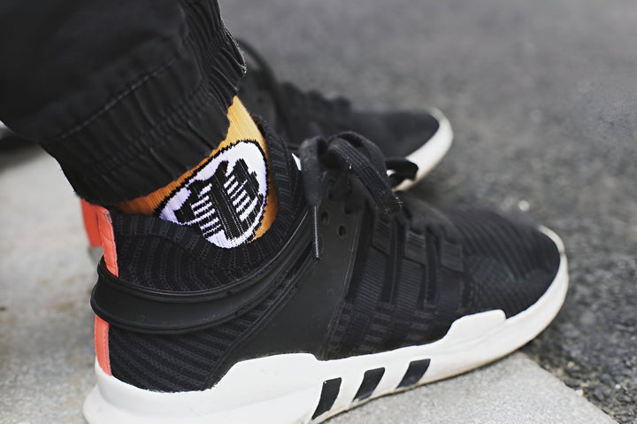 Une personne porte des chaussettes noires avec un motif orange et blanc de Son Goku, un personnage de Dragon Ball Z. Elle porte également des chaussures de sport noires et grises.,CH-0935396012