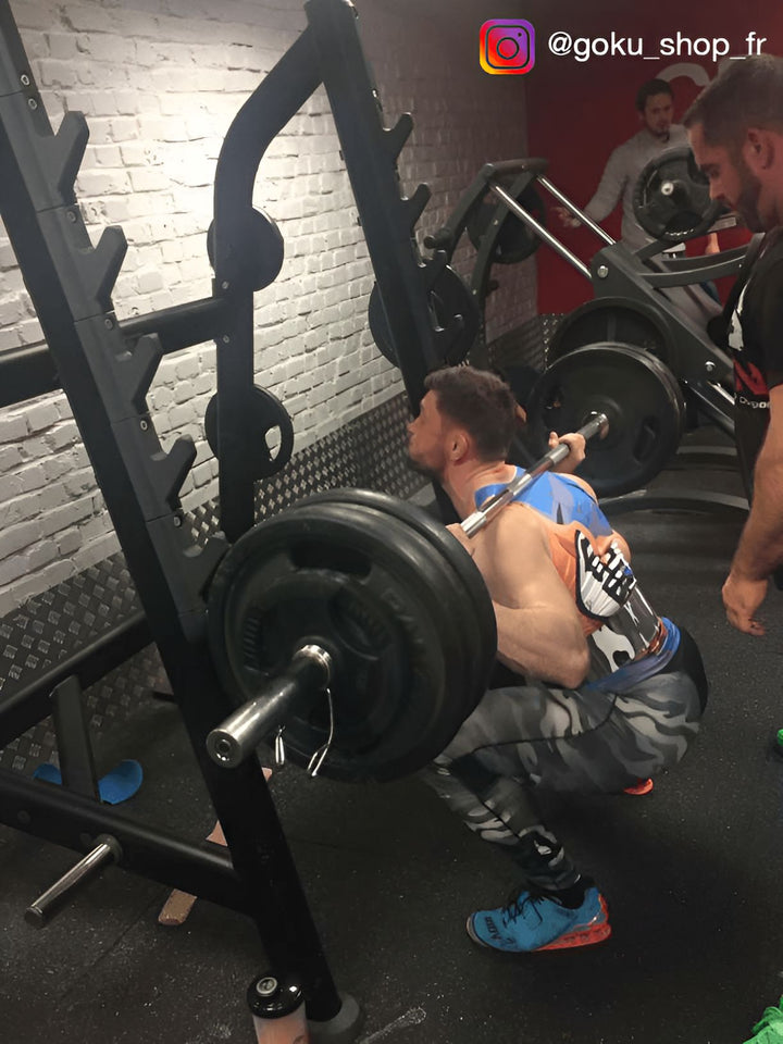 Un homme est en train de faire des squats avec une barre sur les épaules. Il est vêtu d'un débardeur bleu et d'un legging noir. Il est en train de descendre lentement en gardant le dos bien droit.,DS-102924880X,DS-0246837772,DS-1412378486,DS-172070340X,DS-2462884068,DS-5627603399