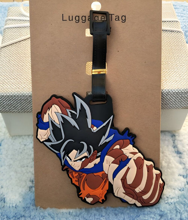 L'image représente un porte-étiquette de bagage en caoutchouc à l'effigie de Son Goku, un personnage de la série animée Dragon Ball Z. Il est vêtu de son costume de combat orange et bleu et est en position de combat. Le porte-étiquette est noir et possède une boucle en métal doré.,É-466797365X