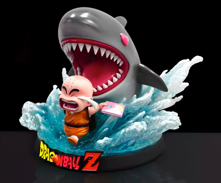 Une figurine de Krilin, un personnage de Dragon Ball Z, en train de se faire attaquer par un requin. Krilin est terrifié et tient un livre à la main. Le requin a la gueule ouverte et est sur le point de l'attaquer. La figurine est posée sur un socle transparent qui représente l'eau.,FI-090087757X