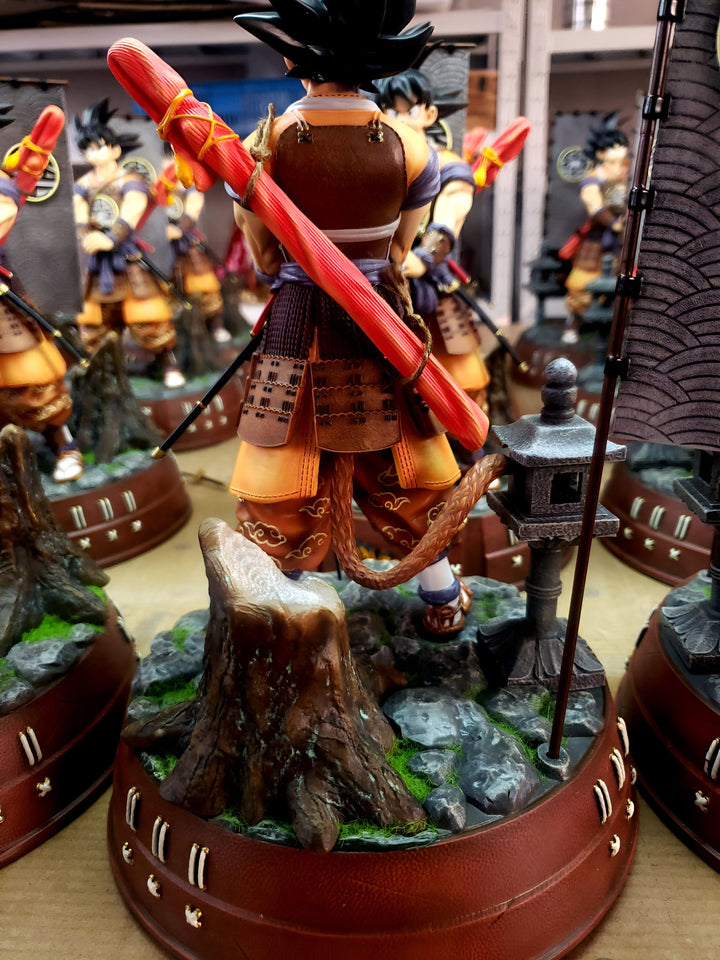 Une figurine de Son Goku, un personnage de la série Dragon Ball Z, est debout sur un rocher. Il porte une armure de samouraï et tient un katana dans sa main droite. Il a une longue queue de singe et des cheveux noirs hérissés. La figurine est très détaillée et mesure environ 30 cm de haut.,FI-1944569502