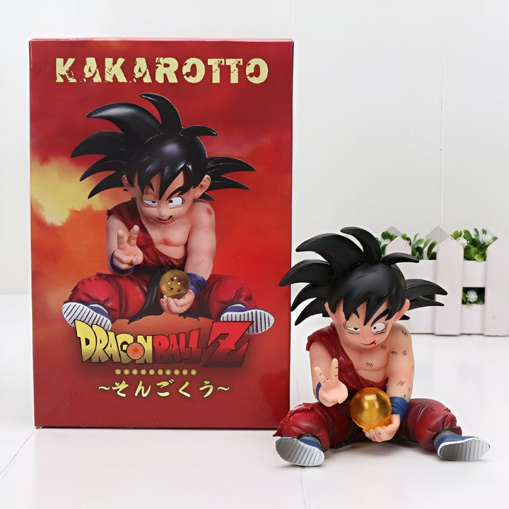 Une figurine de Son Goku, un personnage de Dragon Ball Z, est assise sur une boîte rouge et blanche. Il porte un pantalon orange déchiré et une chemise bleue déchirée. Il a les cheveux noirs et les yeux bleus. Il tient une boule de cristal dans sa main droite.,FI-4324096406