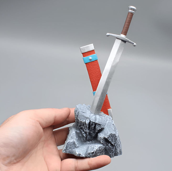 Une épée en métal avec une poignée marron et une lame argentée est posée sur un socle en pierre. La lame de l'épée est légèrement incurvée et la poignée est décorée de deux petites pierres bleues. Le socle en pierre est gris et a une forme de rocher.,FI-7573487673