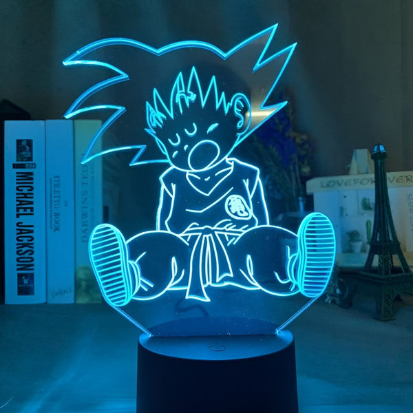 Une image de Son Goku, un personnage de la série animée Dragon Ball Z. Il est assis sur un nuage, les yeux fermés et la bouche ouverte. Il porte son costume orange et bleu. La lumière bleue de la lampe le fait ressortir de l'arrière-plan sombre.,LA-712754834X