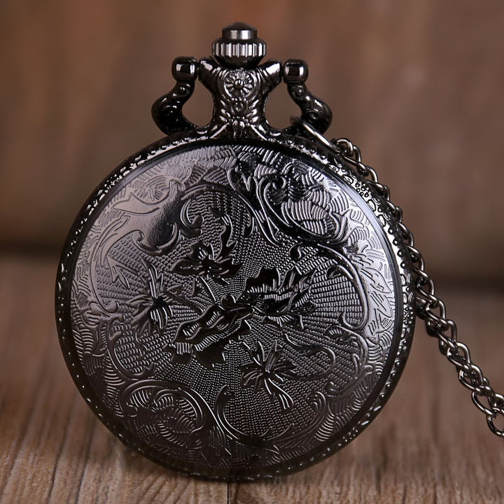 Une image d'une montre de poche noire avec un motif floral sur le couvercle. La montre est ouverte et on peut voir le cadran blanc avec des chiffres romains noirs. La montre est suspendue à une chaîne en métal noir.,MO-5657086066