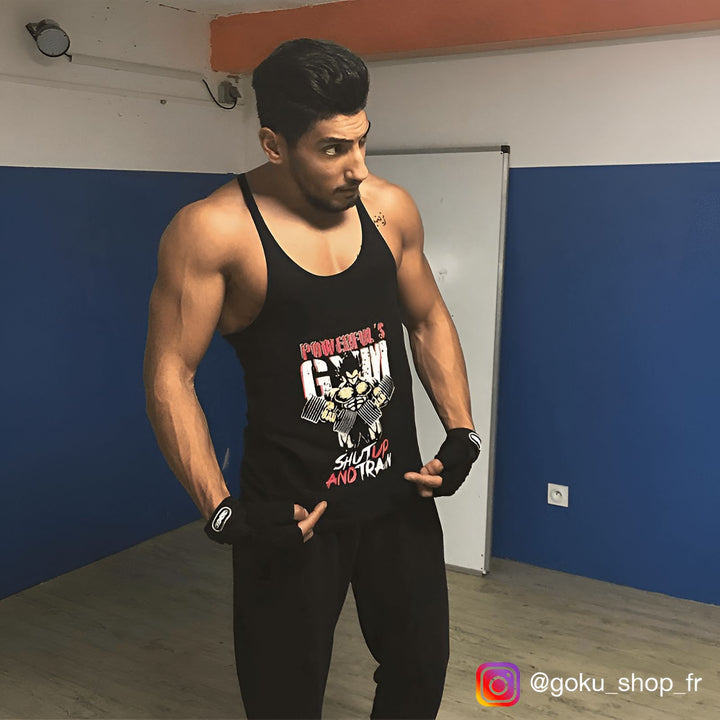 Un jeune homme est vêtu d'un débardeur noir avec un motif coloré représentant le personnage de Son Goku. Il porte également un pantalon de survêtement noir et des gants de musculation. Il se tient debout dans une salle de sport, avec un mur bleu en arrière-plan.,ST-7043545811,ST-135006307X,ST-204086265X