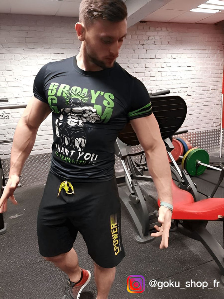 Un homme est en train de poser dans une salle de sport. Il porte un t-shirt noir avec un motif vert, un short noir et des baskets rouges. Il a les cheveux bruns et les yeux marrons. Il est en train de regarder l'objectif de l'appareil photo.,T--7078037022,T--6329442959,T--8295411284,T--4536798889,T--0188599355