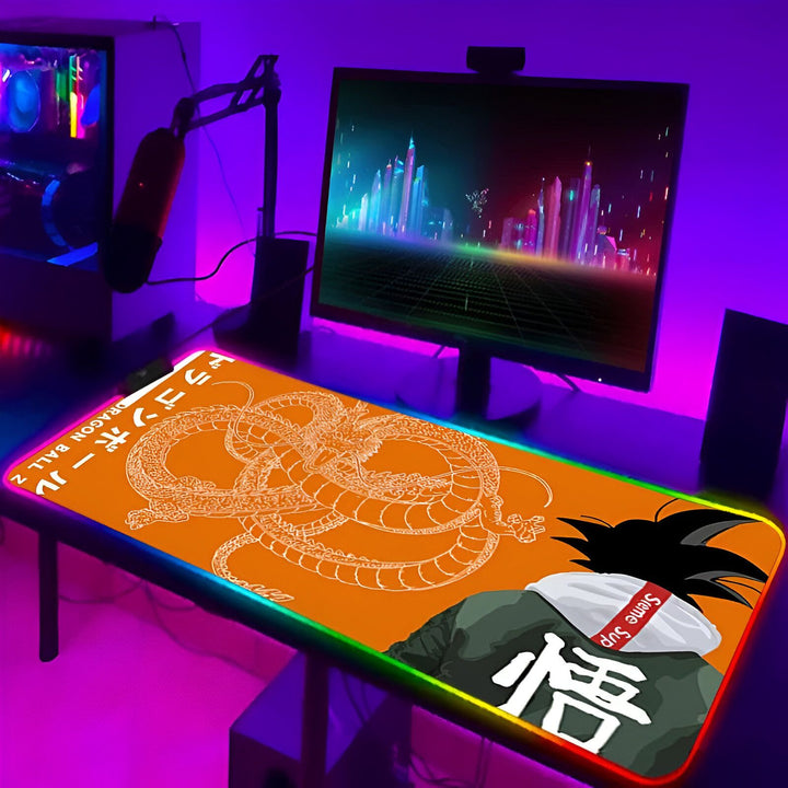 L'image représente un tapis de souris sur un bureau de gamer. Le tapis de souris est orange et noir avec un personnage de Dragon Ball Z dessus. Il y a un écran d'ordinateur, un clavier et une souris sur le bureau. L'arrière-plan est un mur avec une lumière violette.,TA-2375721535,TA-455495479X,TA-4045812474